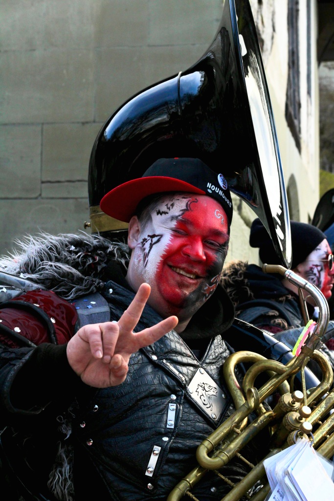 A man in a parade with a face painted red and white and carrying a trombone. 
